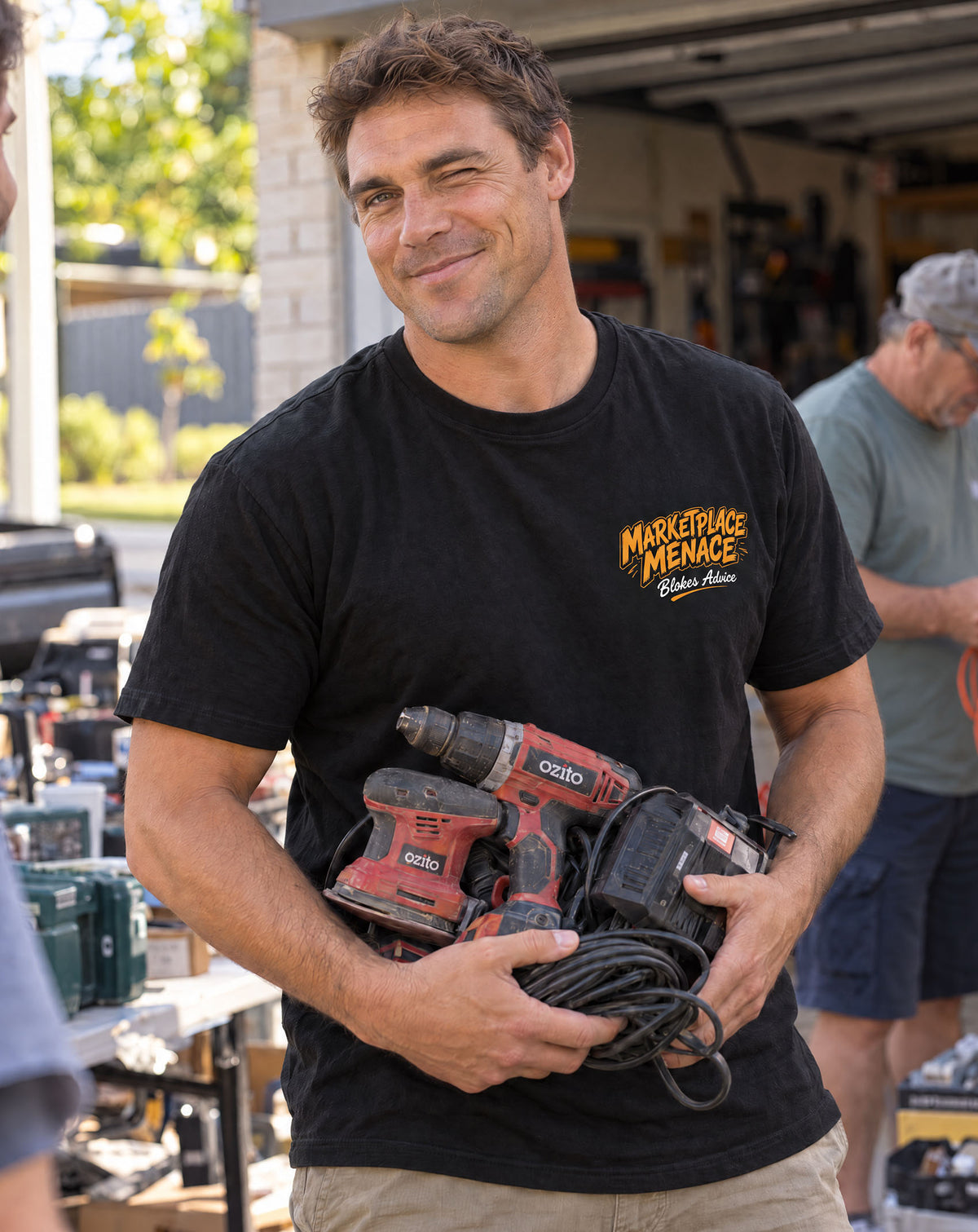 Bloke grinning in the black Marketplace Menace Tee with orange chest logo, holding a cordless drill at a garage sale